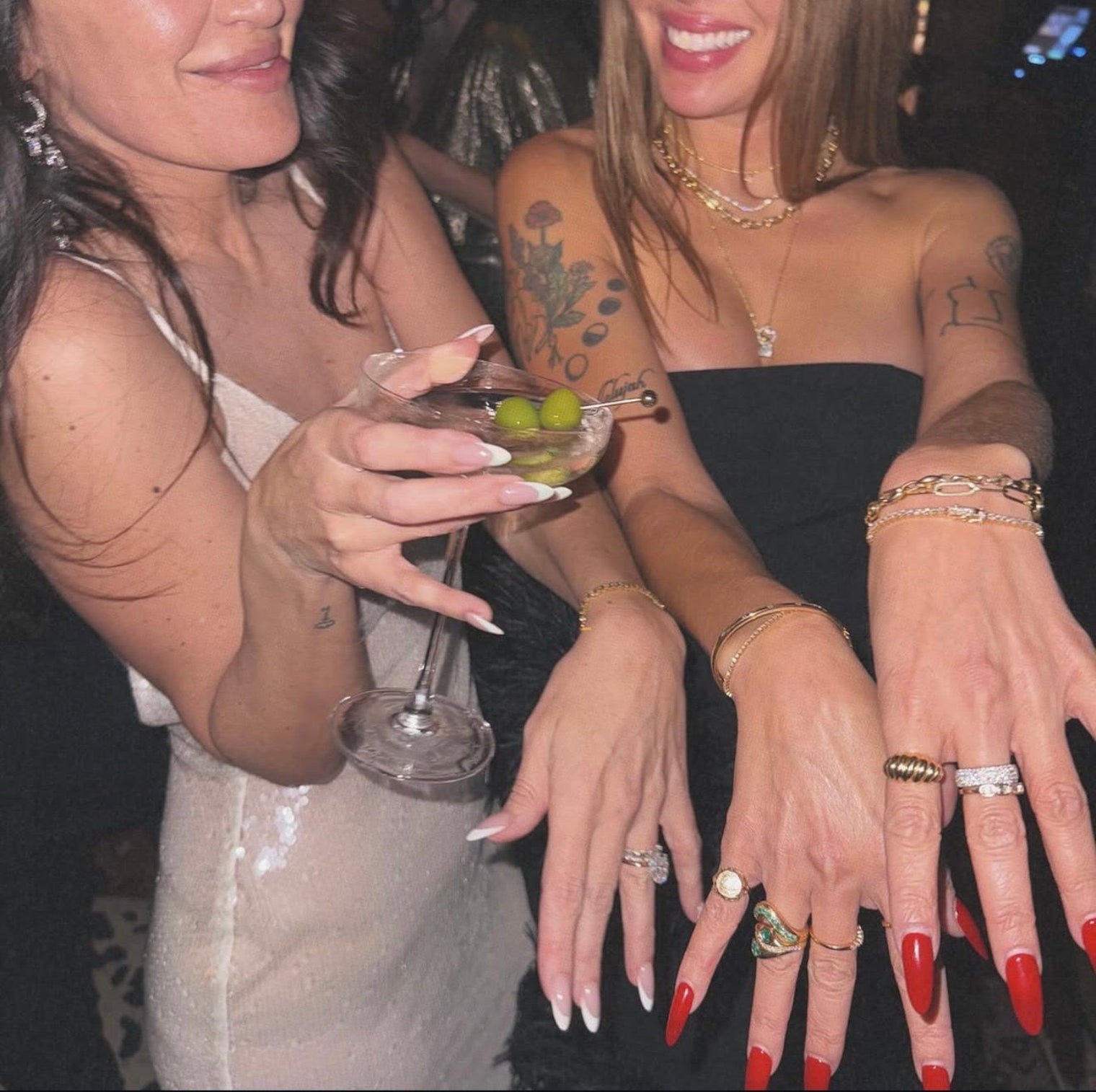 Two women at a social event, one holding a cocktail, with tattoos and jewelry on display.